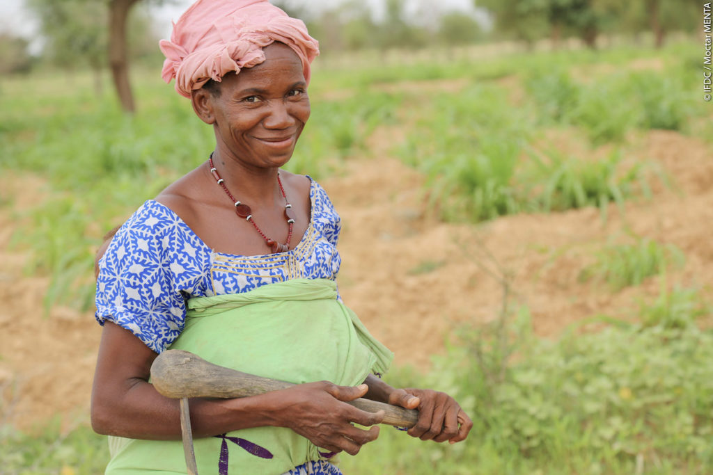 A woman stands in a field in Mali with a farming hoe over her shoulder.