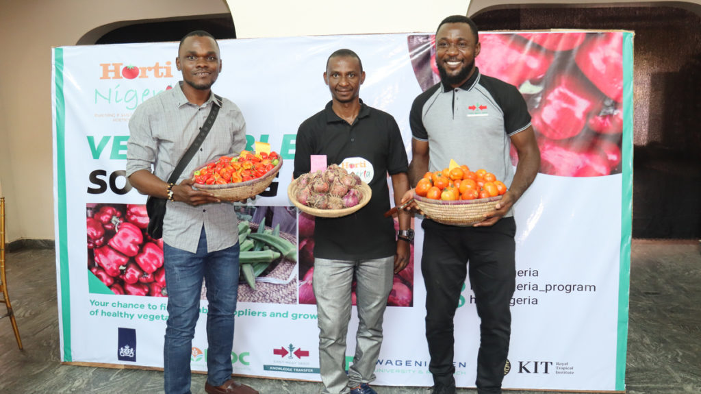 Three male HortiNigeria program partners pose with bowls of their produce — peppers, onions, and tomatoes.