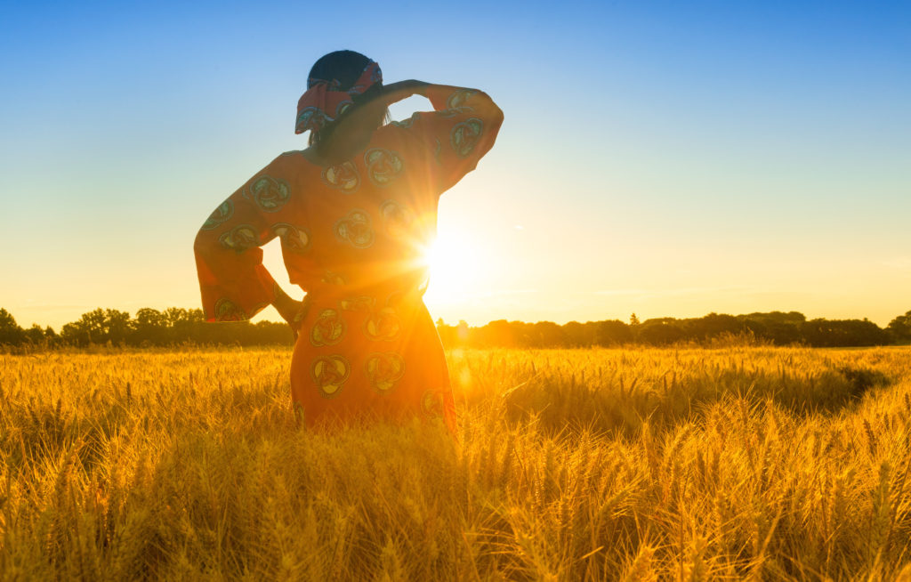 African woman in traditional clothes standing, looking, hand to eyes, in field of barley or wheat crops at sunset or sunrise