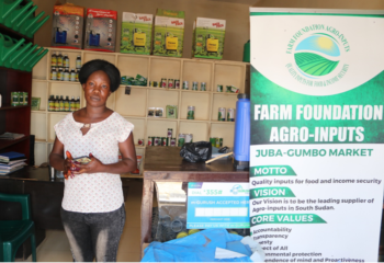 Agrodealer Clara Keji, a South Sudanese woman in a white shirt and blue jeans, stands to the left of a large sign in her shop