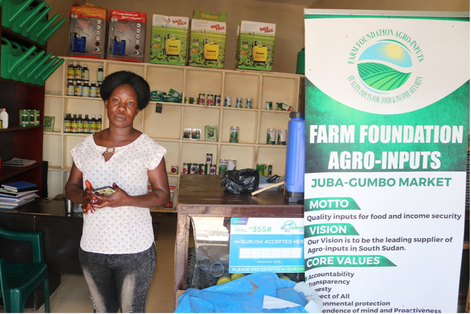 Agrodealer Clara Keji, a South Sudanese woman in a white shirt and blue jeans, stands to the left of a large sign in her shop