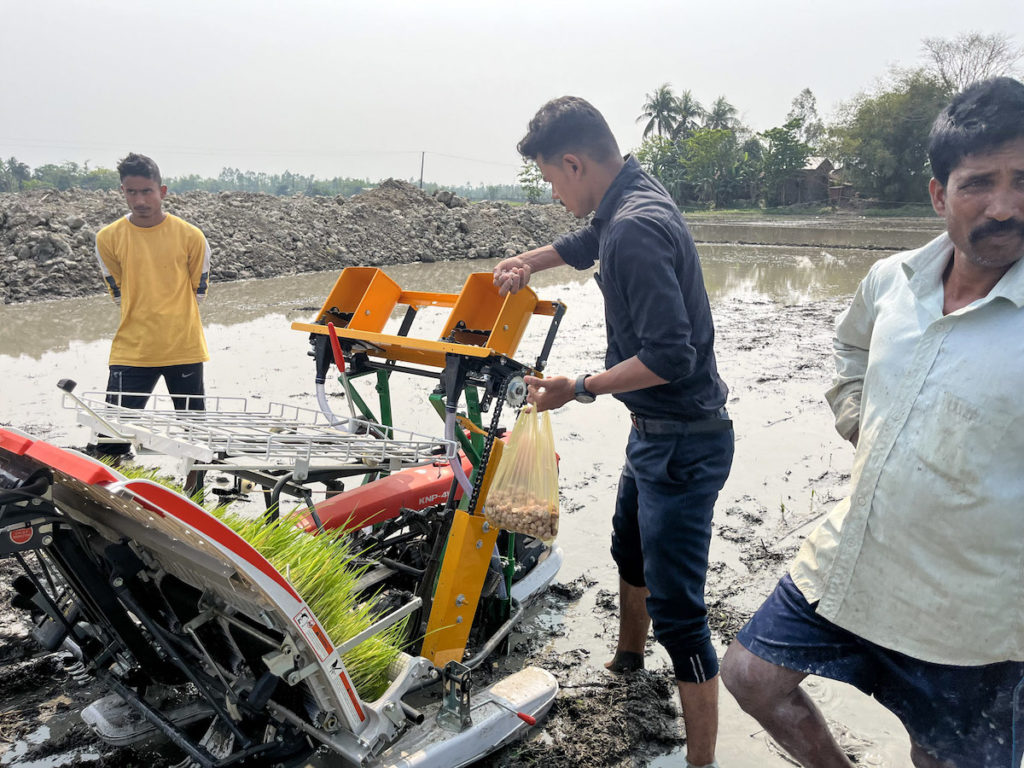 Men use a seed-and-fertilizer drill to apply UPK briquettes in a flooded rice field