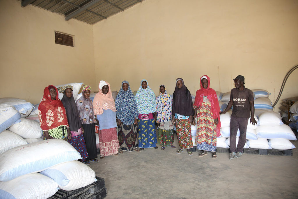 Eight women and one man stand in a warehouse with large bags surrounding them