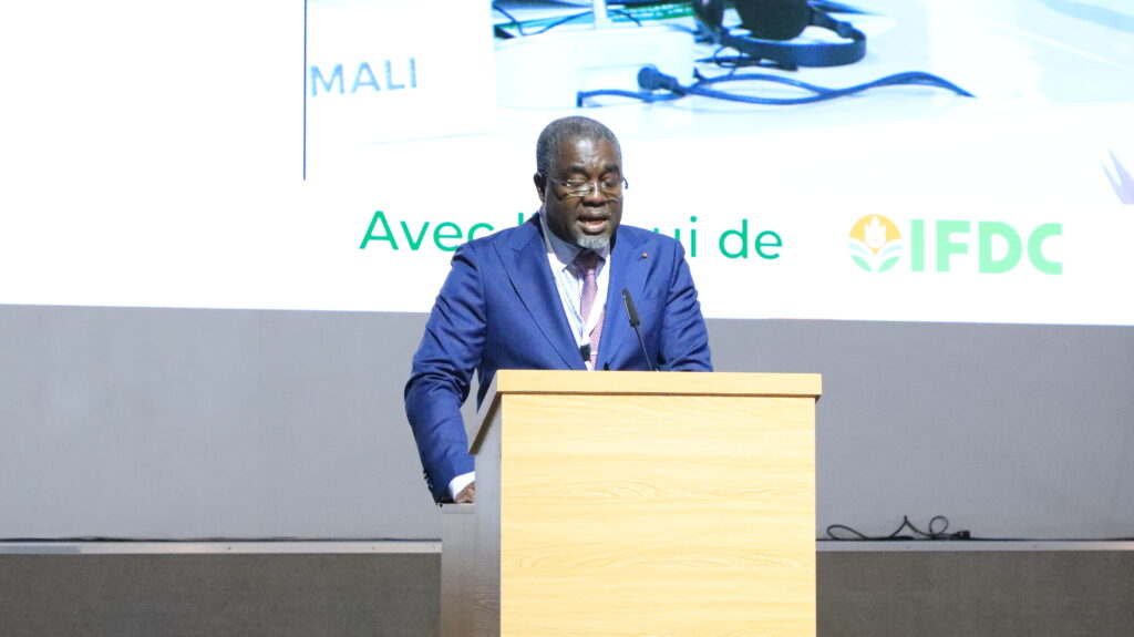 A speaker in a blue suit with a pink tie stands at a wooden podium