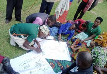 A group of people gathered around outside on the ground and standing as someone draws on a big sheet of paper about their vision.
