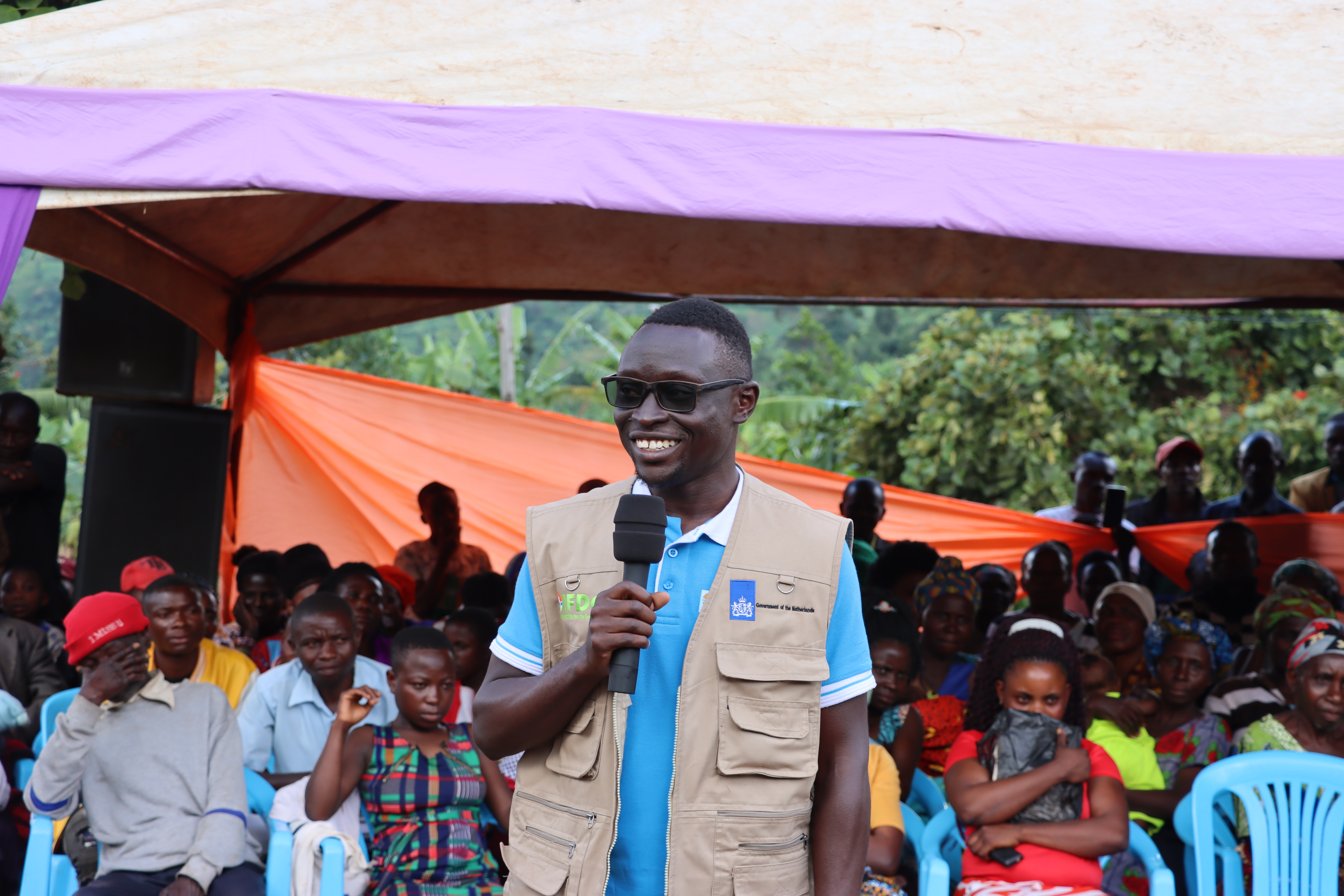 A man stands with a microphone in front of a group of people under a purple tent