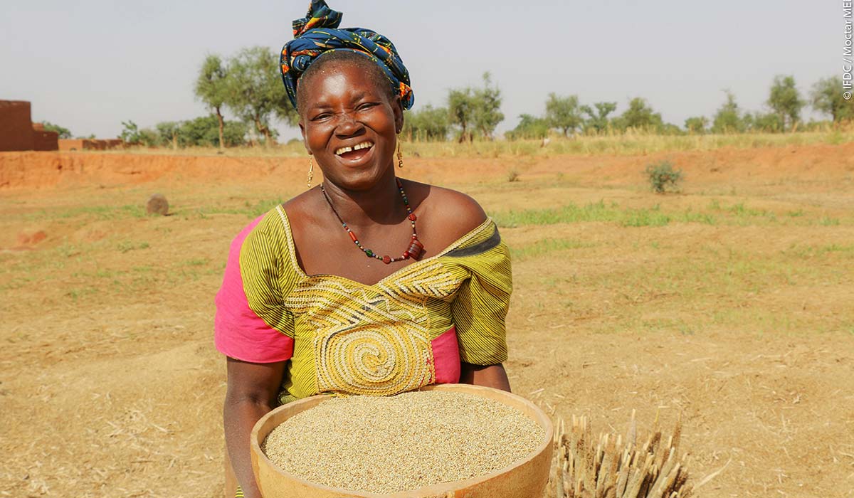 Nana Guido holds a basket of Sorghum and smiles.